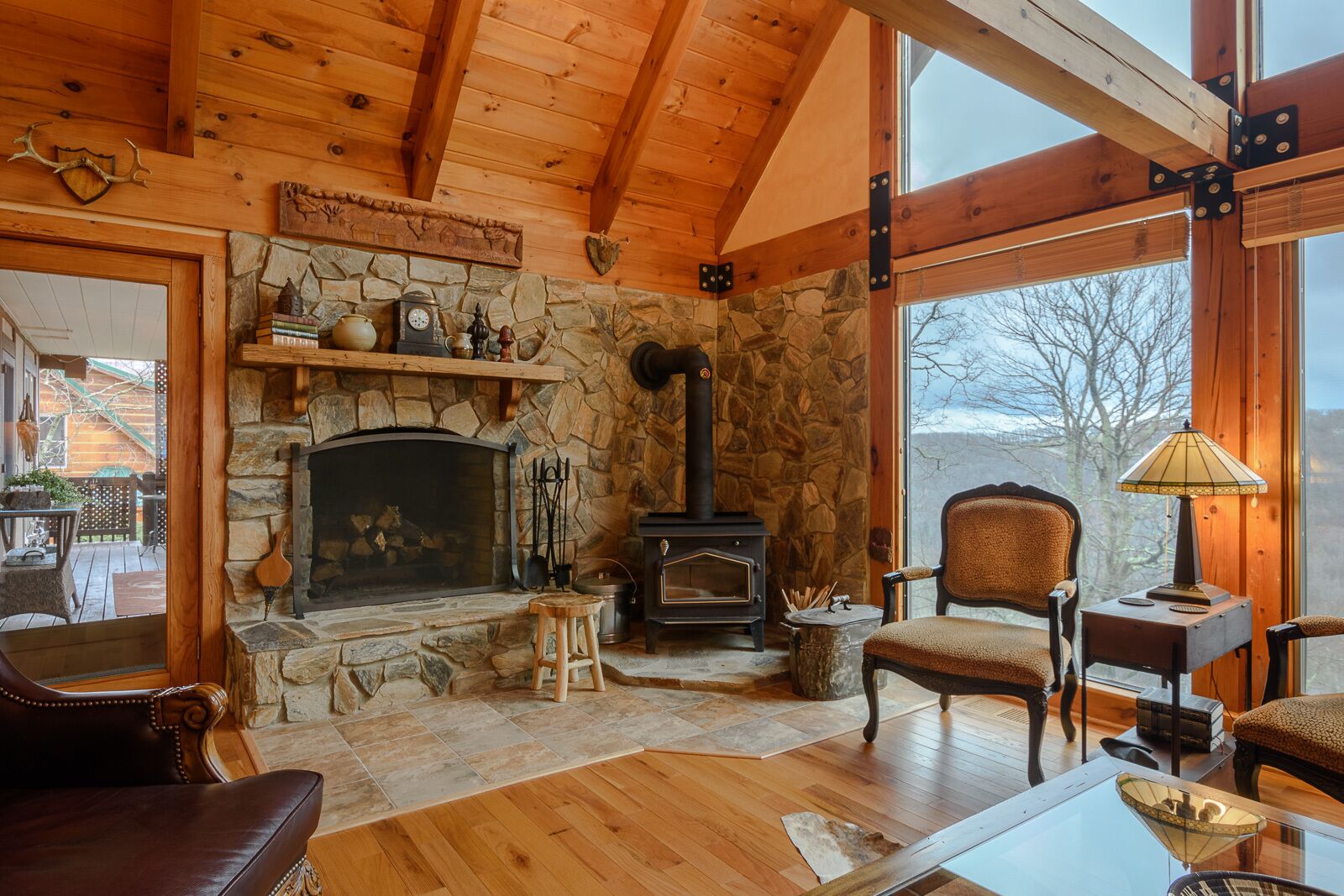 A cozy cabin interior with a stone fireplace, wooden beams, and a large window offering a view of the outdoors.