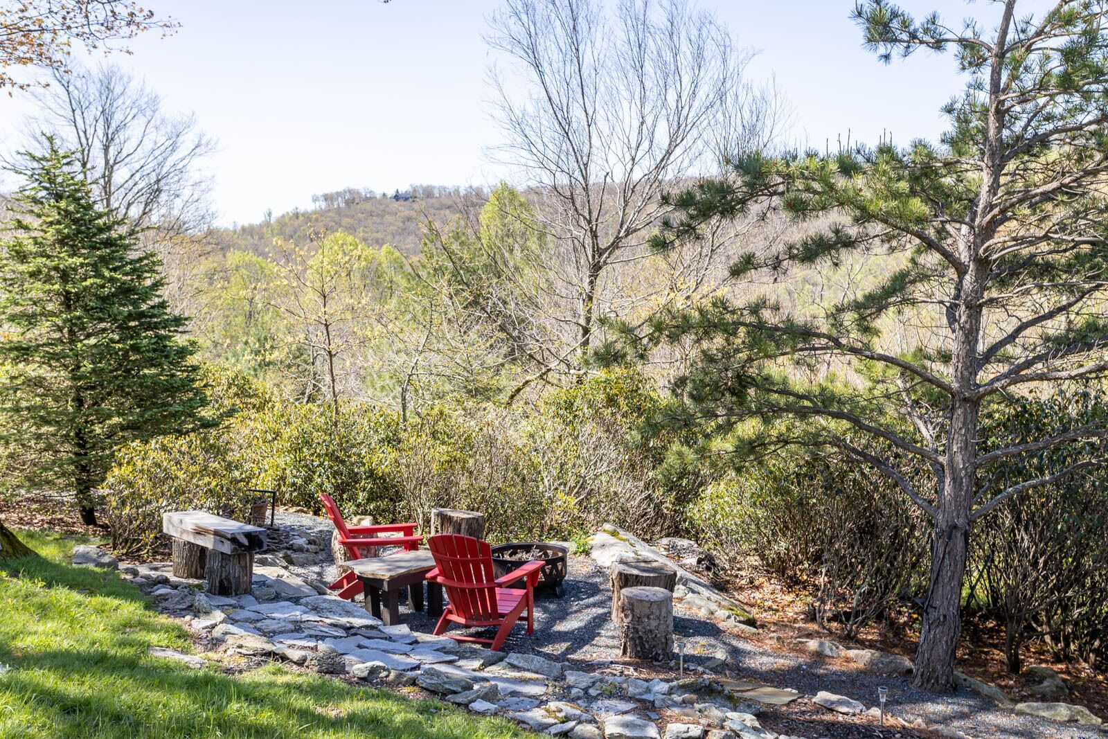 An outdoor seating area with a stone table and red chairs, surrounded by trees and a hillside in the background.