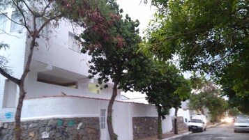 Un edificio blanco con base de piedra, árboles en primer plano y coches aparcados en la calle.