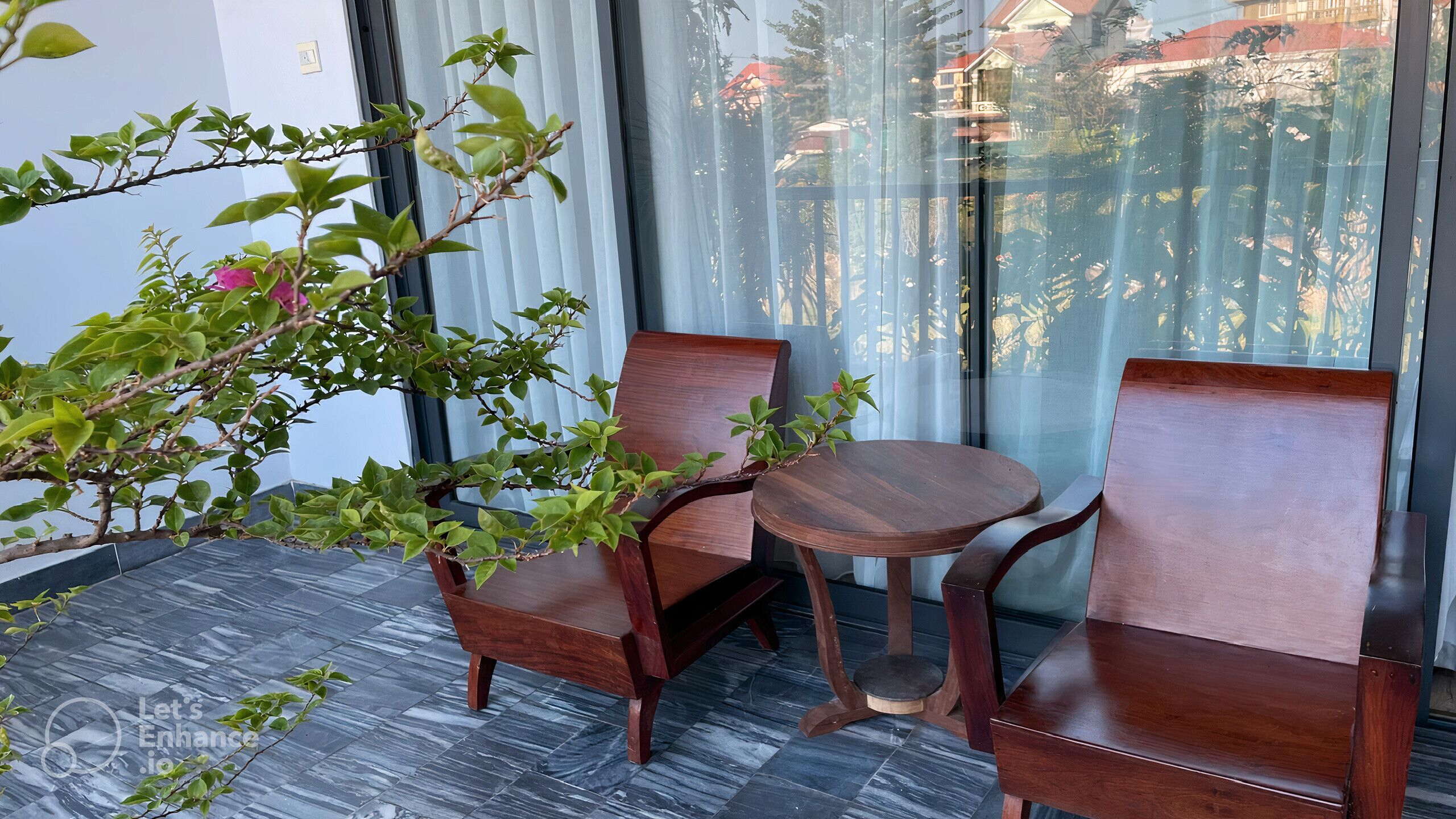 A small, modern room with a dark tiled floor, two wooden chairs, a small round table, and a potted plant with green leaves and a pink flower.