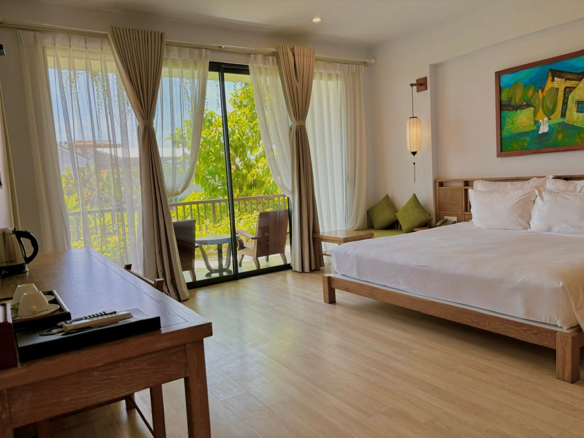 A hotel room with a large bed, a desk with a kettle, a chair, and a balcony with curtains.