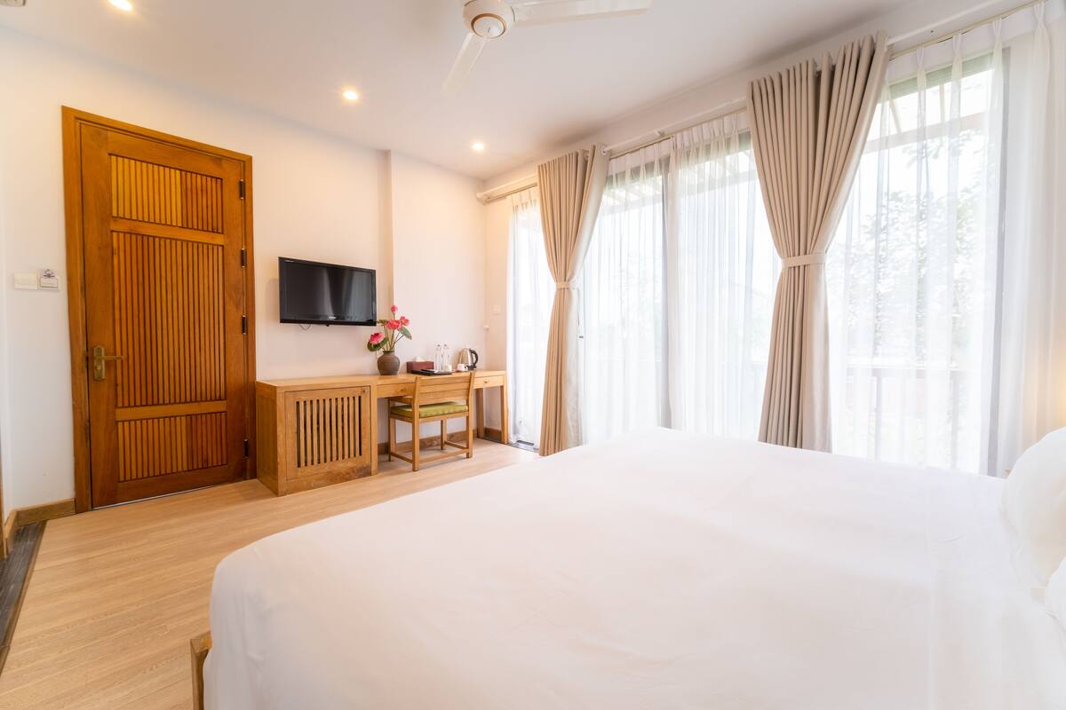 Une chambre d’hôtel avec un grand lit, un bureau en bois avec une chaise, un téléviseur fixé au mur et une fenêtre avec des rideaux.