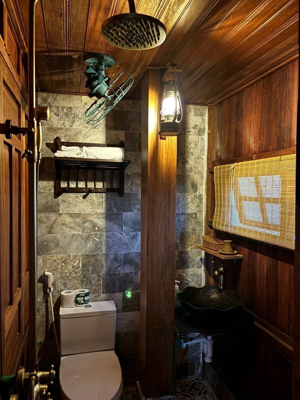 A compact bathroom with a toilet, a shelf with towels, a hanging shower head, and a wall-mounted light fixture.