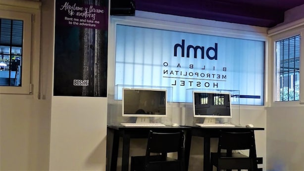 Une chambre d’hôtel équipée de deux bureaux avec ordinateur, chacun comprenant un écran et un clavier. Une grande affiche sur le mur porte les inscriptions « nmd » et « BIBLIOTECOL ELECTRONIC HOTEL ».