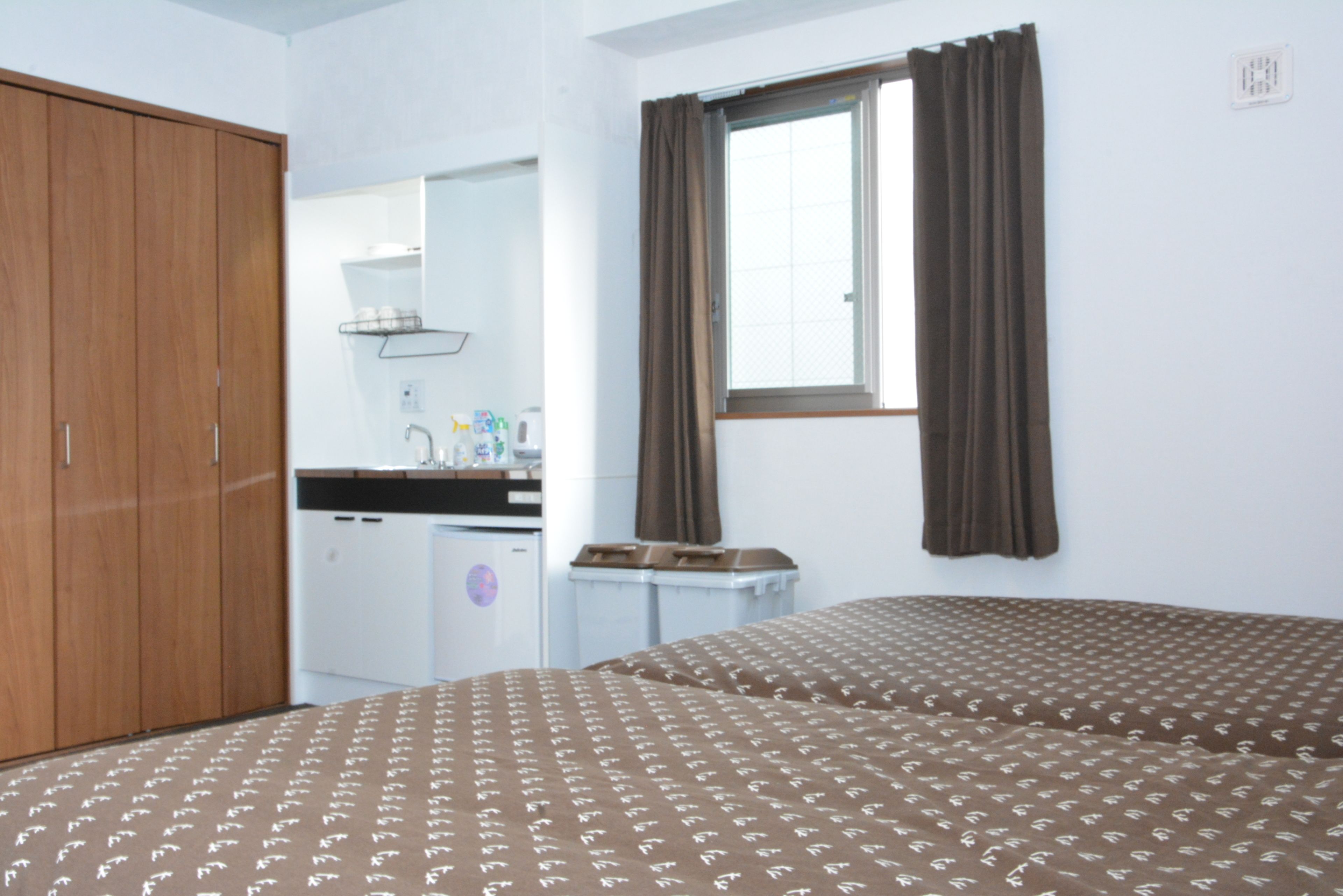 A room with two beds, a wooden wardrobe, a small kitchenette, and a window with curtains.