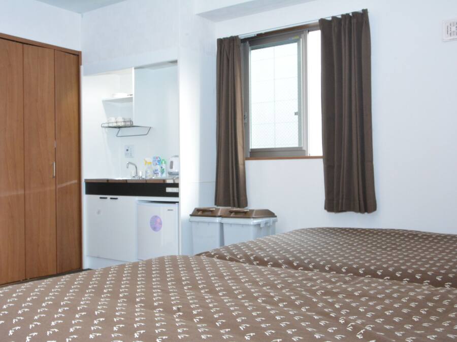 A room with two beds, a wooden wardrobe, a small kitchenette, and a window with curtains.