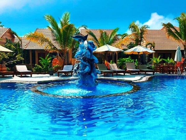 A swimming pool with a central fountain, surrounded by lounge chairs and umbrellas, with palm trees and a building in the background. - Golden Topaz Resort (Phu Quoc)