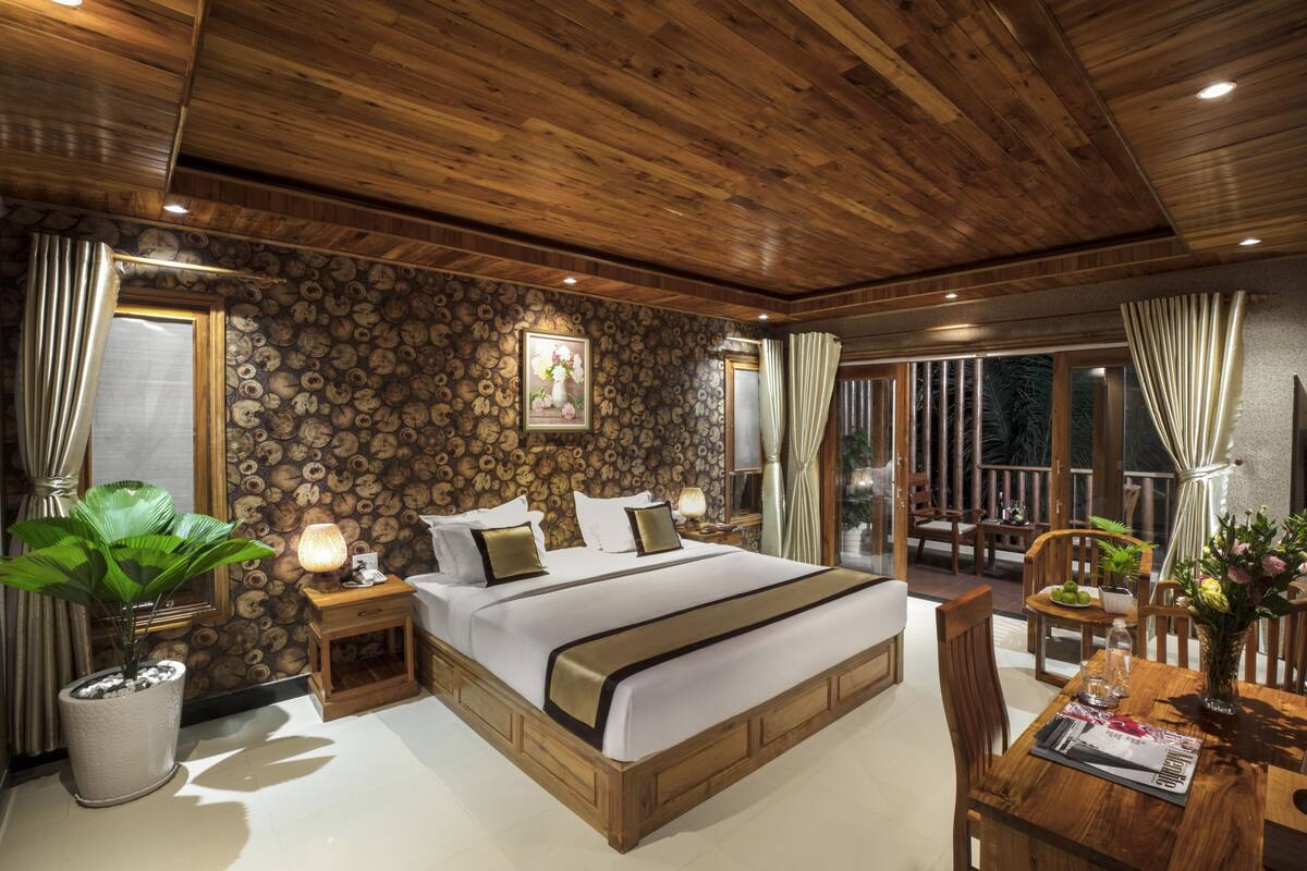 Une chambre avec un plafond en bois, un grand lit avec une tête de lit, une table de chevet avec une lampe, une plante en pot et une table à manger avec des chaises.