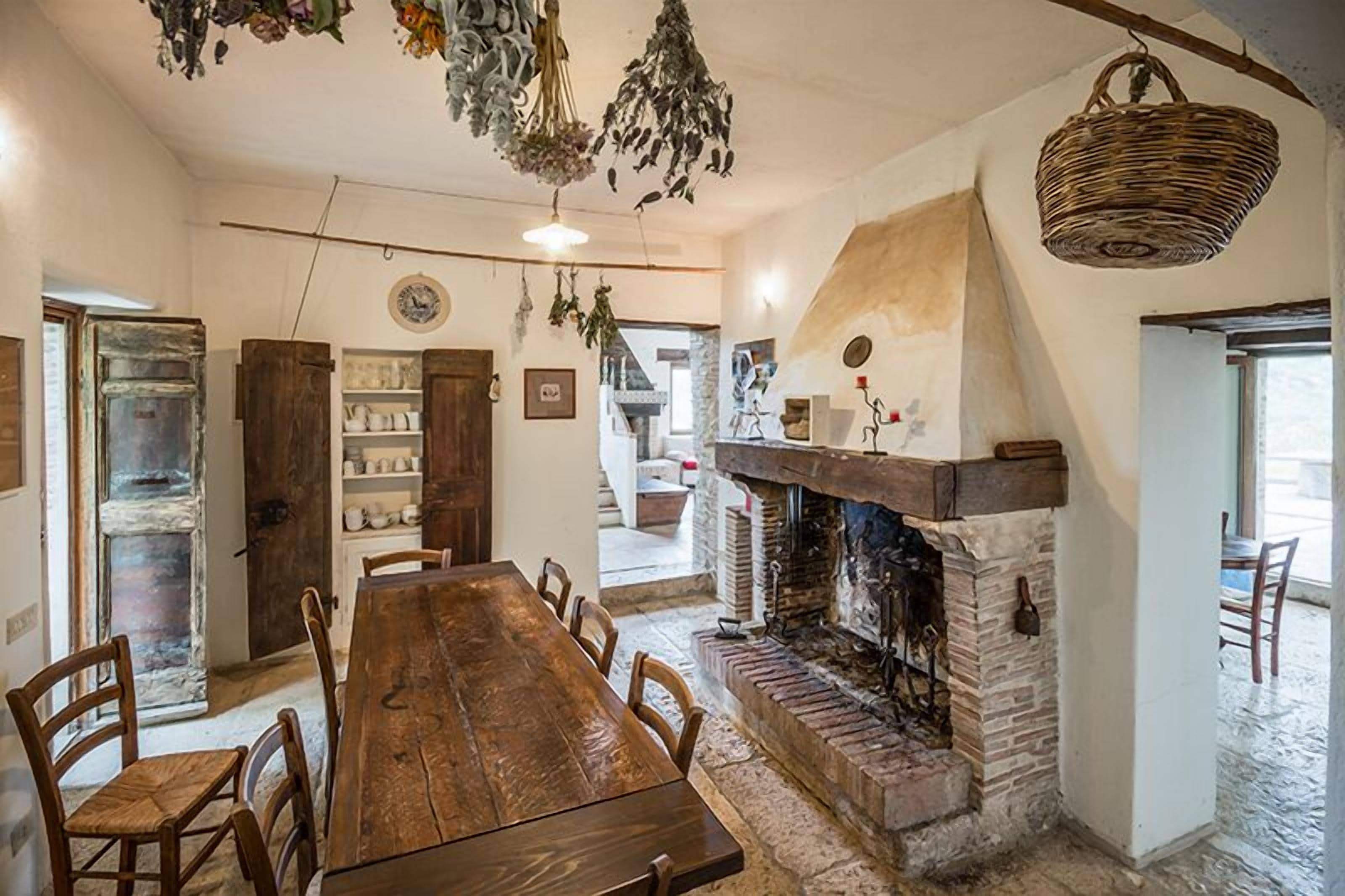 A rustic dining area with a long wooden table and chairs, a stone fireplace, and hanging plants.
