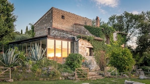 Una casa de piedra con un invernadero de cristal, rodeada por una piscina y abundante vegetación.