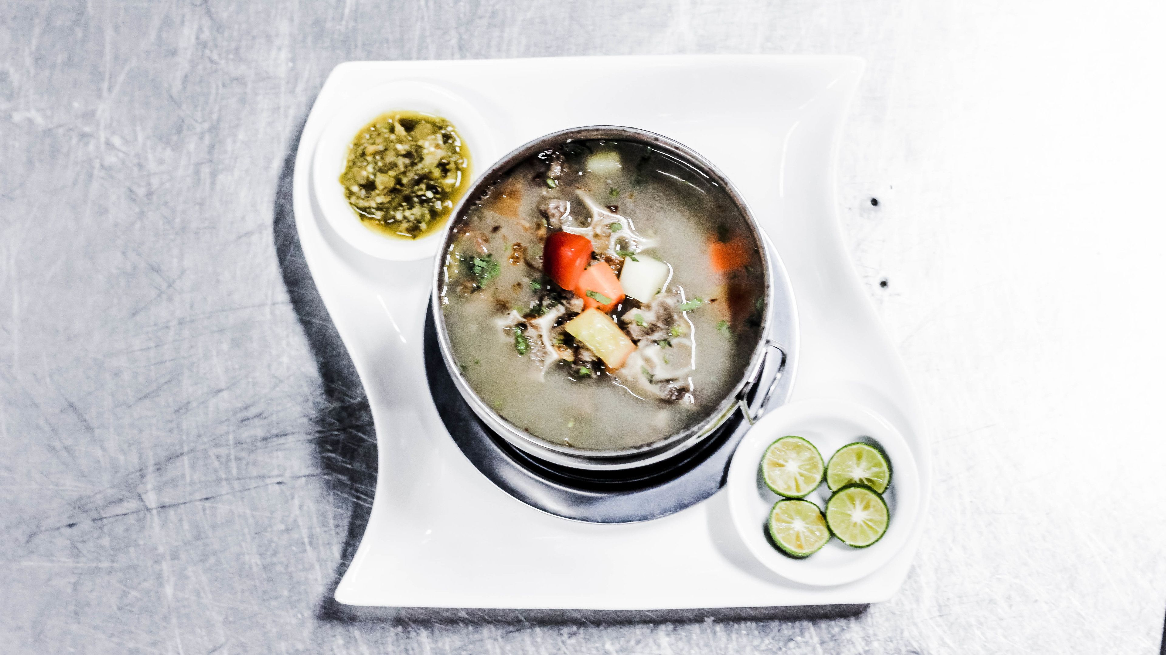Un tazón de sopa con verduras y carne, acompañado de un recipiente pequeño de salsa y unas cuñas de lima sobre un plato blanco.