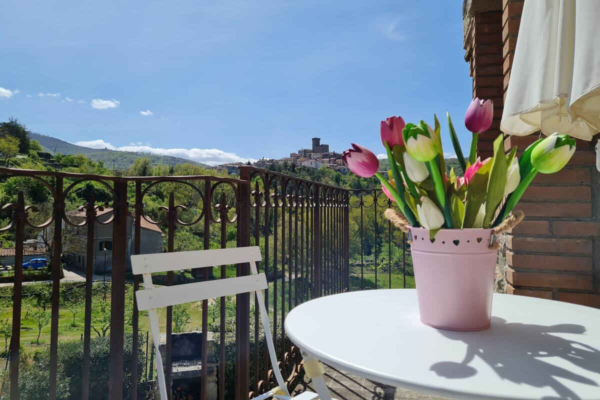 Un balcón con una mesa blanca y una maceta rosa de tulipanes, con vistas a un paisaje verde y un pueblo lejano.