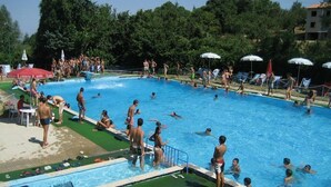 Una piscina con gente en el agua y en la solana, rodeada de árboles y sombrillas.