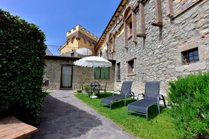 Un bâtiment en pierre avec un chemin pavé, un espace extérieur aménagé avec une table et des chaises, et un parasol blanc.