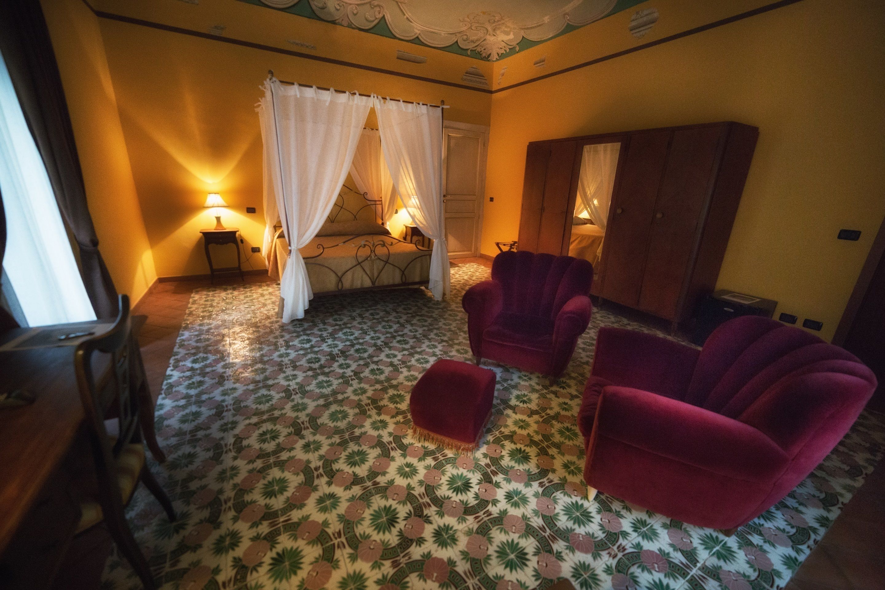Una habitación con una cama, dos sillones rojos, un armario de madera y un suelo estampado.