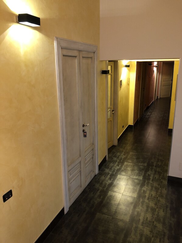 Un couloir d’hôtel avec un mur de couleur claire, un revêtement de sol foncé et des portes au-dessus desquelles sont fixés des luminaires.