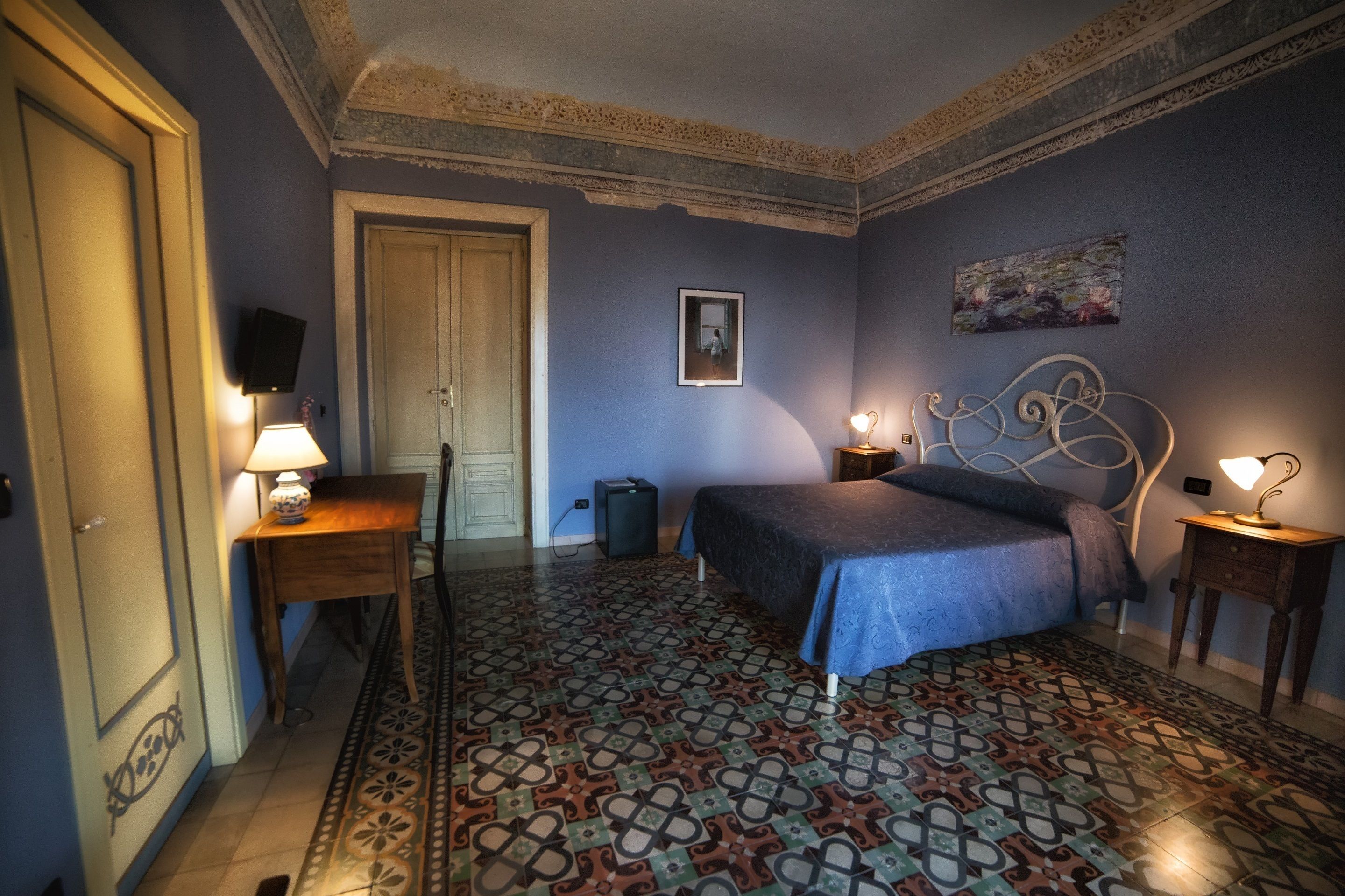 Un dormitorio con piso estampado, una cama con cabecera, un escritorio con lámpara y una puerta.