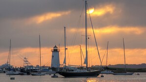 A harbor with multiple boats docked, a prominent lighthouse, and a sunset in the background. - Pristine Chappy! August 29 through Labor Day 2026 Available !  (Chappaquiddick Island, Edgartown)