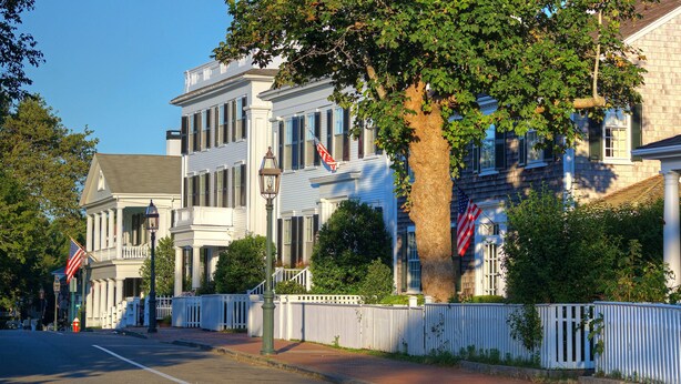 A row of historic buildings with traditional architecture, a white picket fence, and American flags. - Pristine Chappy! August 29 through Labor Day 2026 Available !  (Chappaquiddick Island, Edgartown)