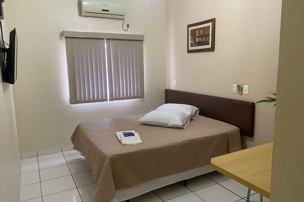 Habitación de hotel con cama, mesita de noche, equipo de aire acondicionado, ventana con persianas, un cuadro colgado en la pared y suelo de baldosas.