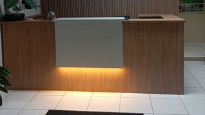 A hotel reception desk with a digital screen above it, a wooden counter, and a reception area with a tiled floor. - Hotel Marcan (Sumare)