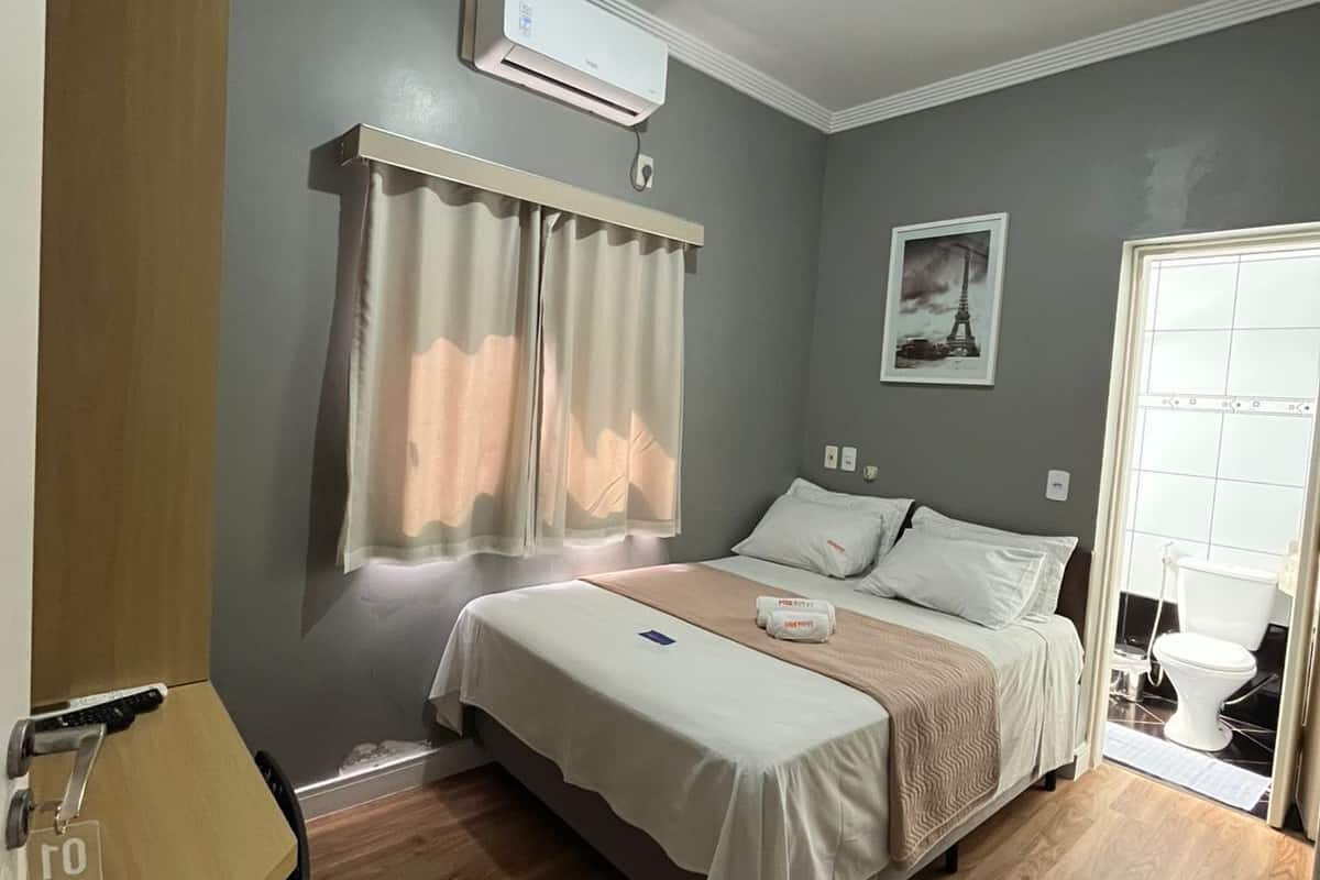 Un dormitorio con cama, cortinas, una lámpara en el techo, aire acondicionado de pared y una puerta que da al baño.