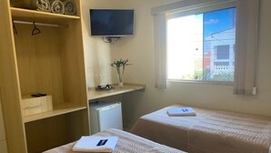 A hotel room with two beds, a TV mounted on the wall, a small desk with a plant, and a window with a view of buildings. - Hotel Marcan (Sumare)