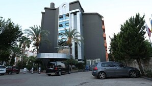 A modern building with a prominent entrance, a parking area with several cars, and palm trees in the background. - Mysea Hotels Incekum (Alanya)