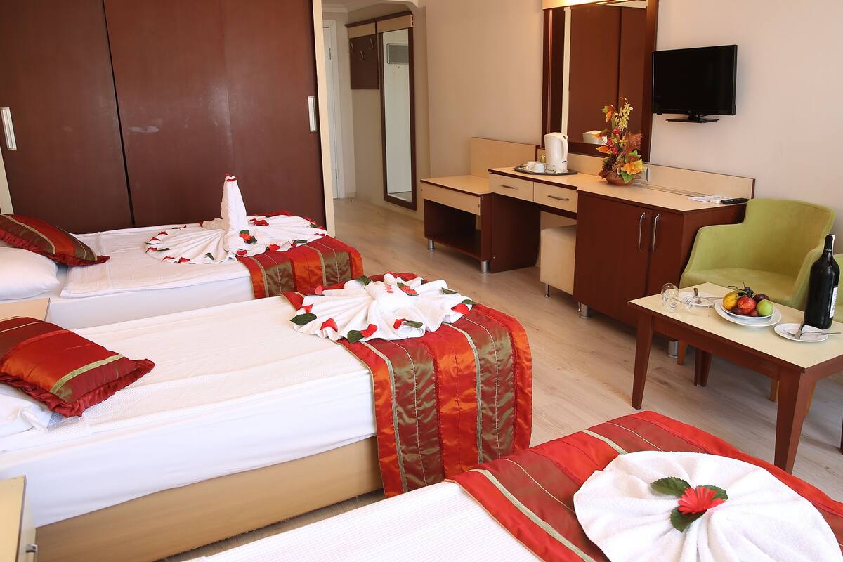 A hotel room with two beds, a desk, a chair, a TV, and a dining table with a bottle and fruit.