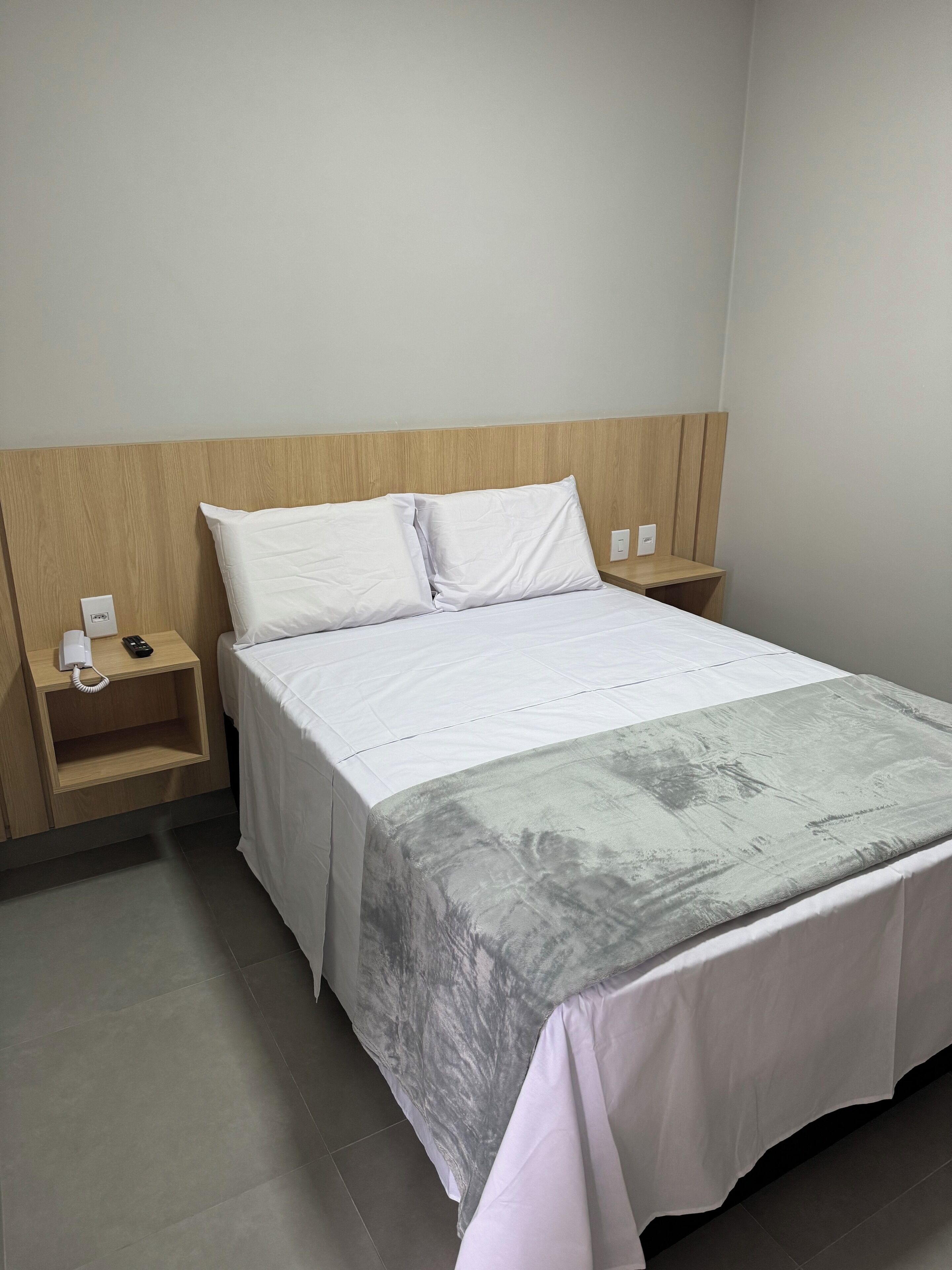 A neatly made bed with white linens and a grey blanket, a wooden headboard, a bedside table with a phone and a power outlet, and a plain wall.