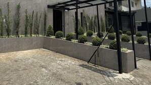 A modern hotel with a sign reading "HS HOTEL". It features a covered entrance with a glass canopy, a paved courtyard, and neatly arranged greenery.