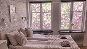 Une chambre avec un grand lit, deux tables de chevet, un radiateur et de grandes fenêtres donnant sur la forêt.