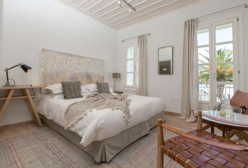 A bedroom with a bed, bedside table, lamp, and a view of the outdoors through a window.