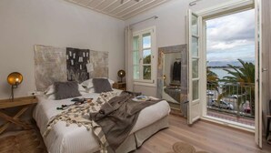 A bedroom with a bed, a nightstand, a wall-mounted lamp, and a view of the outside through an open door. - Amymone Suites (Nafplio)
