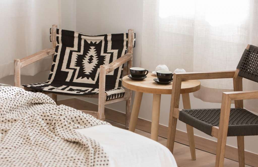 Ein Zimmer mit einem Bett, zwei Holzstühlen, einem kleinen runden Tisch mit zwei Tassen und einem weißen Gegenstand sowie einer gemusterten Decke auf einem Stuhl.