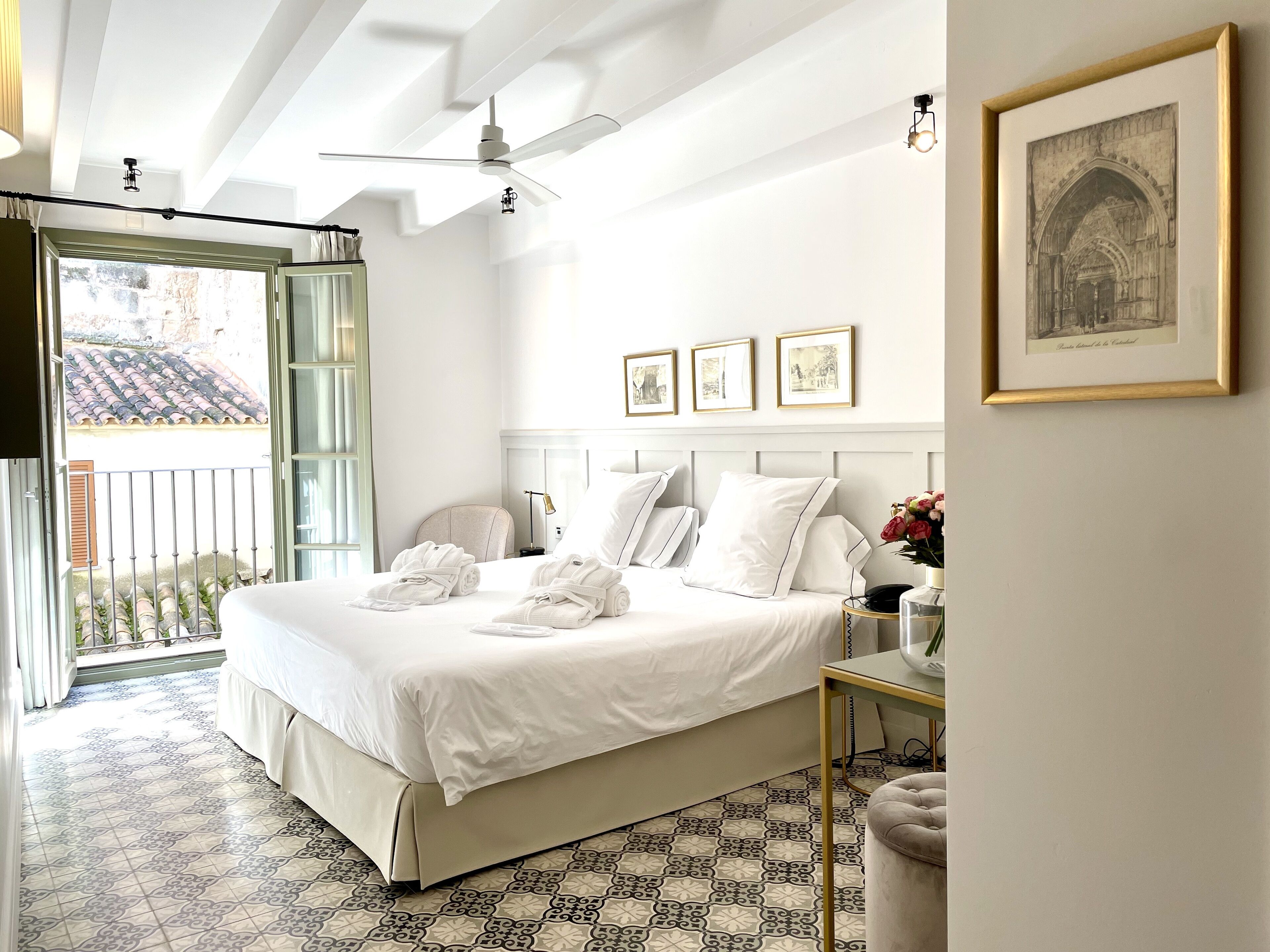 Habitación de hotel con una cama grande, ventilador de techo, balcón con vistas y fotografías enmarcadas en la pared.