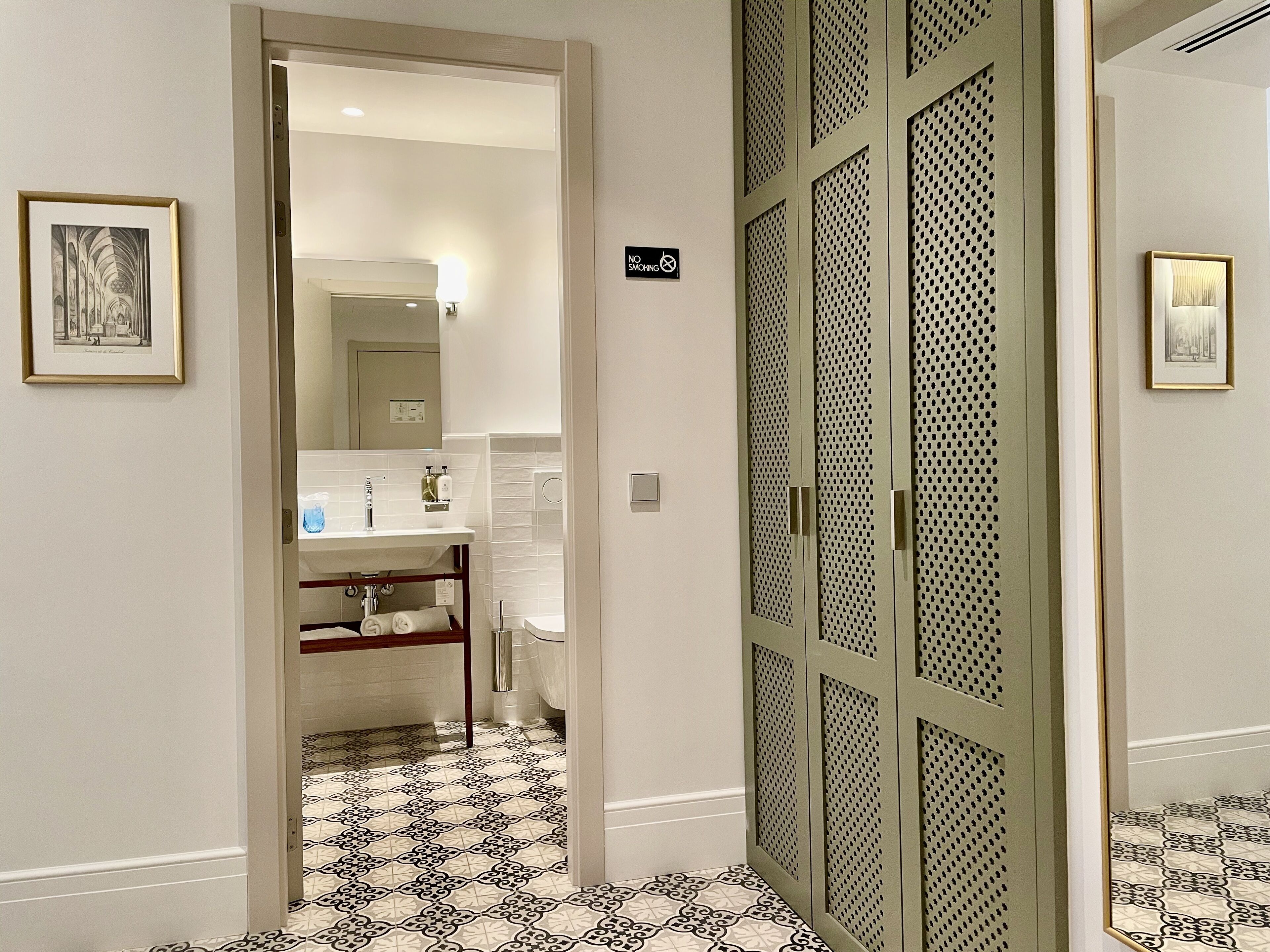 Un bany d'hotel amb un lavabo, mirall i vàter. Hi ha una porta amb un senyal damunt.