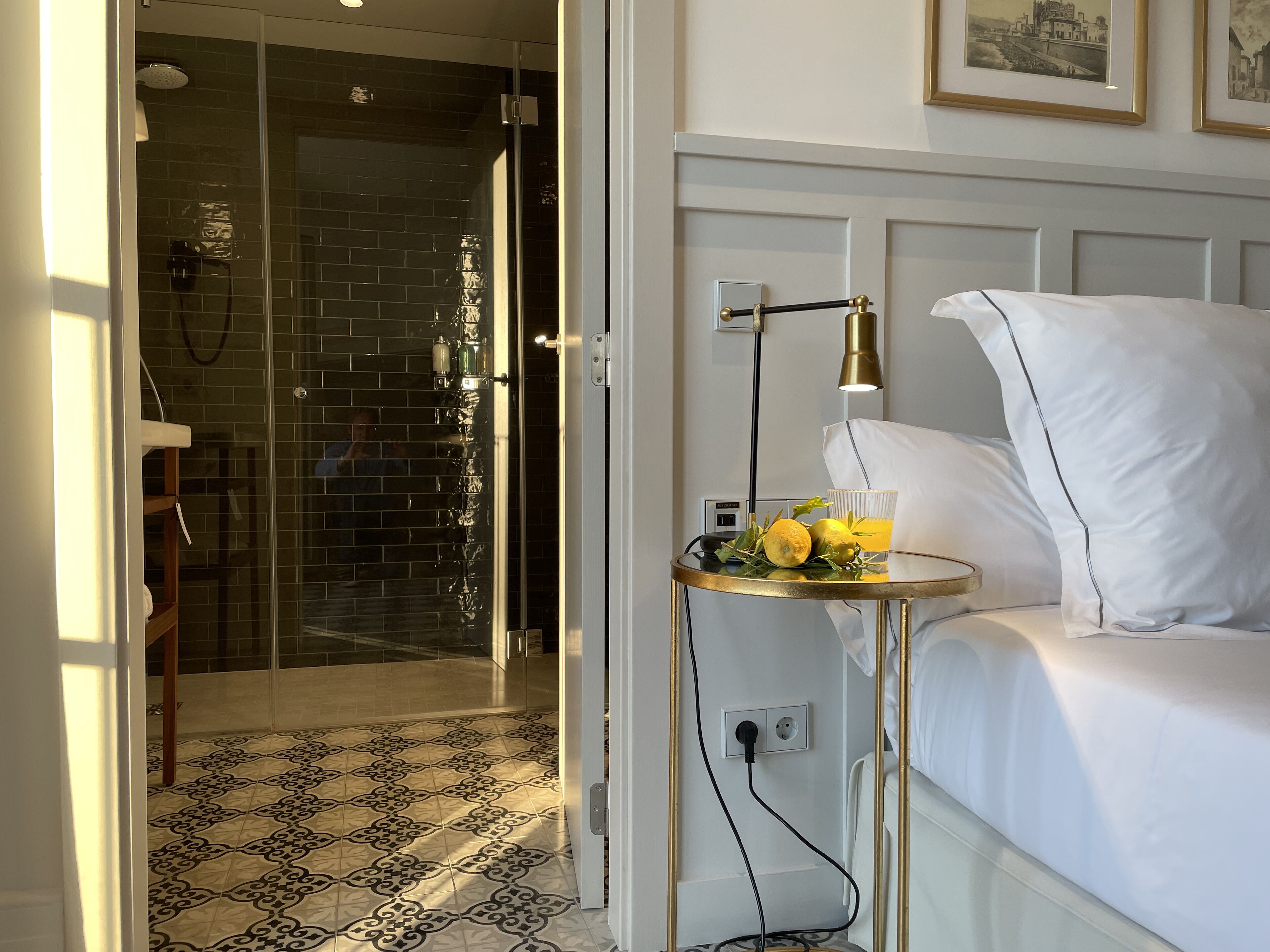 A hotel room with a bed, a bedside table with a lamp, a glass of orange juice, and a shower area with a glass door.