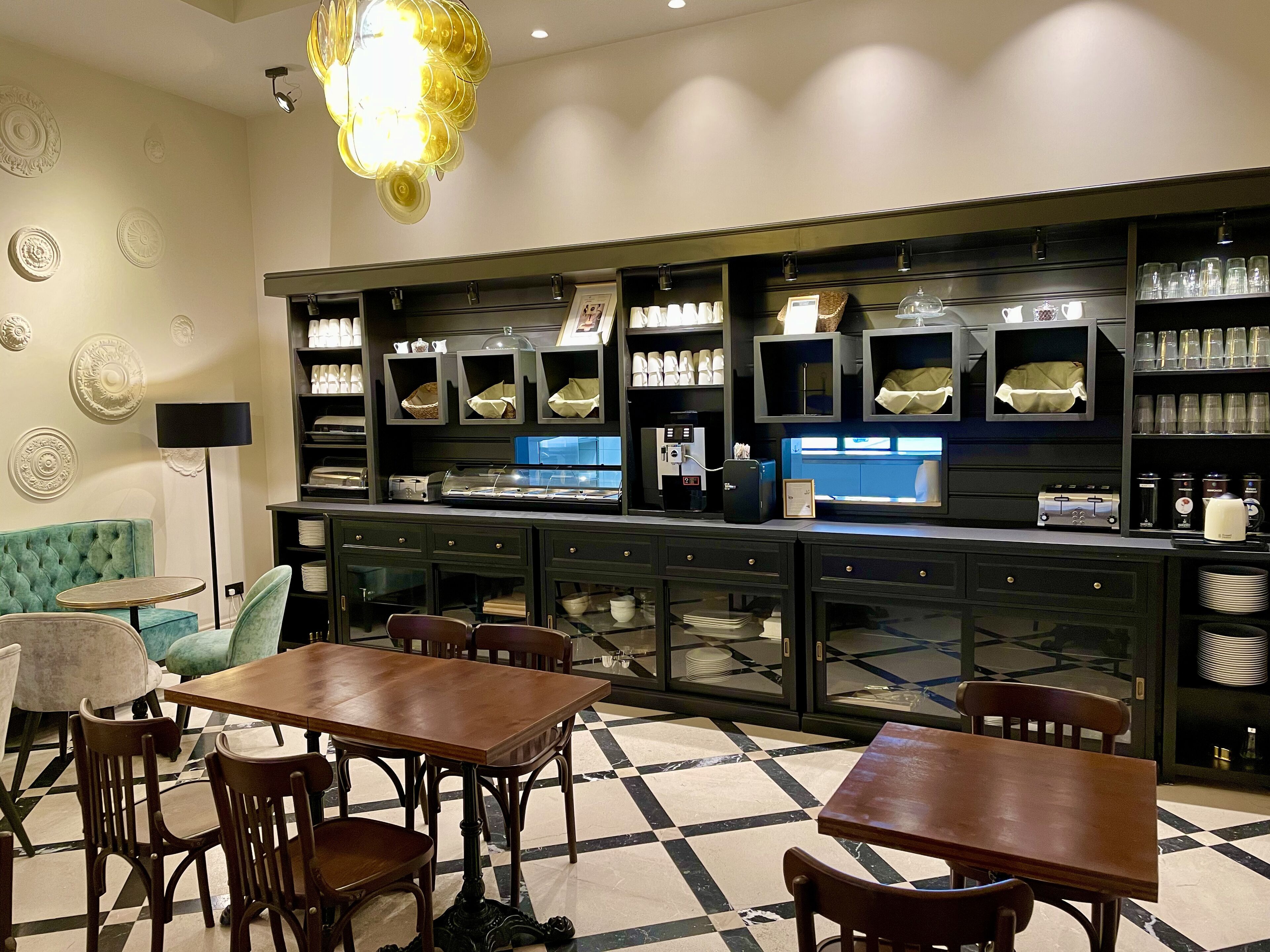 Un restaurante con suelo a cuadros, mesas y sillas de madera. Se ven estanterías con cristalería y una máquina de café.