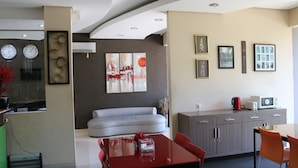 A modern living room with a grey sofa, a red dining table, and a kitchen area with a microwave and kettle. - Tirta Mansion Premier (Curug)