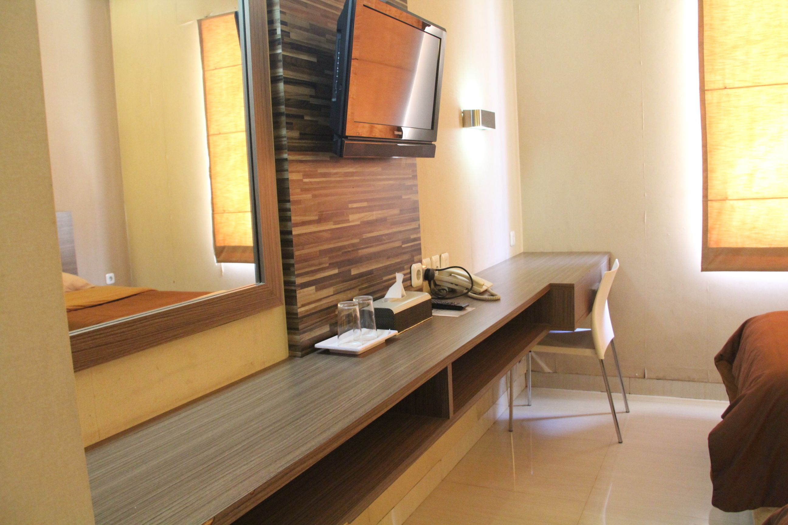 Ein Hotelzimmer mit einem hölzernen Waschtisch, einem Spiegel, einem an der Wand befestigten Flachbildfernseher, einem Schreibtisch mit Telefon, Gläsern und einer Taschentuchbox.