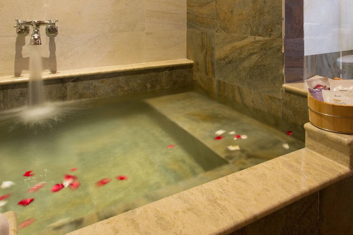 A bathtub with a marble surround, a glass enclosure, and a wooden shelf with a potted plant and a small container.