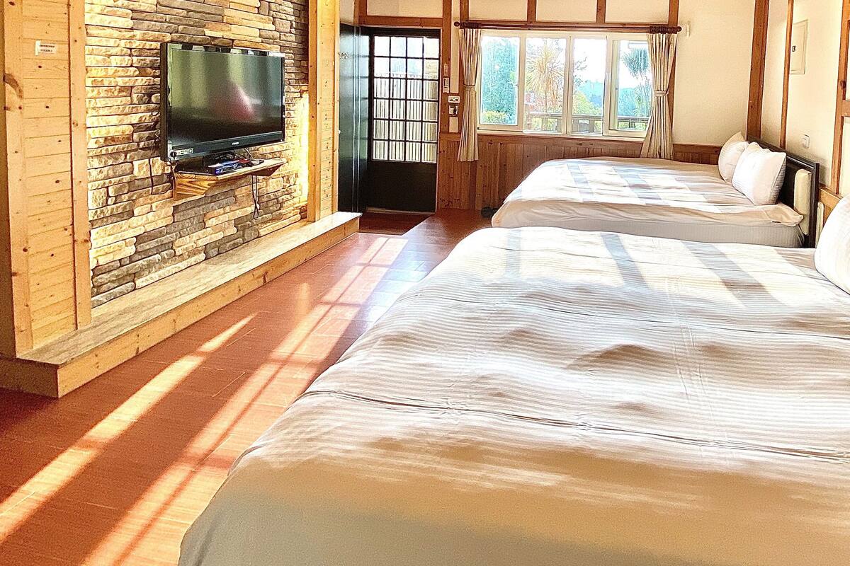 A room with two beds, a TV mounted on a stone wall, and a window with curtains.