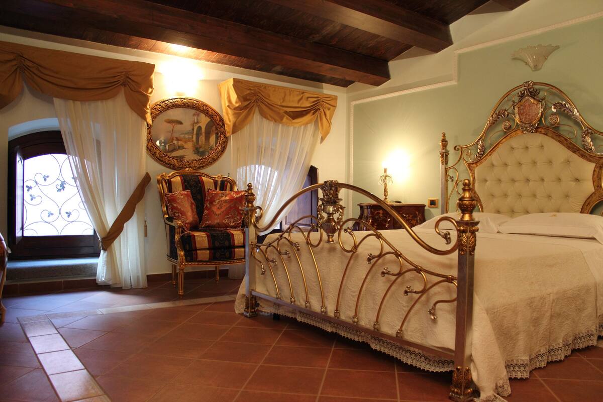 Un dormitorio con una cama con dosel, una silla con cojín, un espejo y una ventana con cortinas.