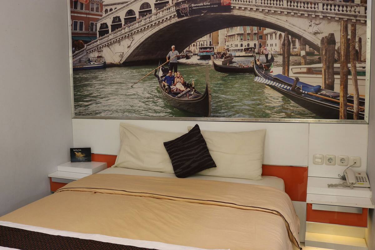 A hotel room with a bed, a nightstand, a lamp, and a wall-mounted painting of a canal scene.