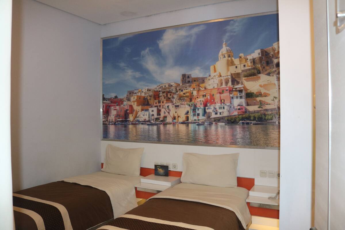 A hotel room with two beds, a large painting of a coastal town, and a view of the sea.