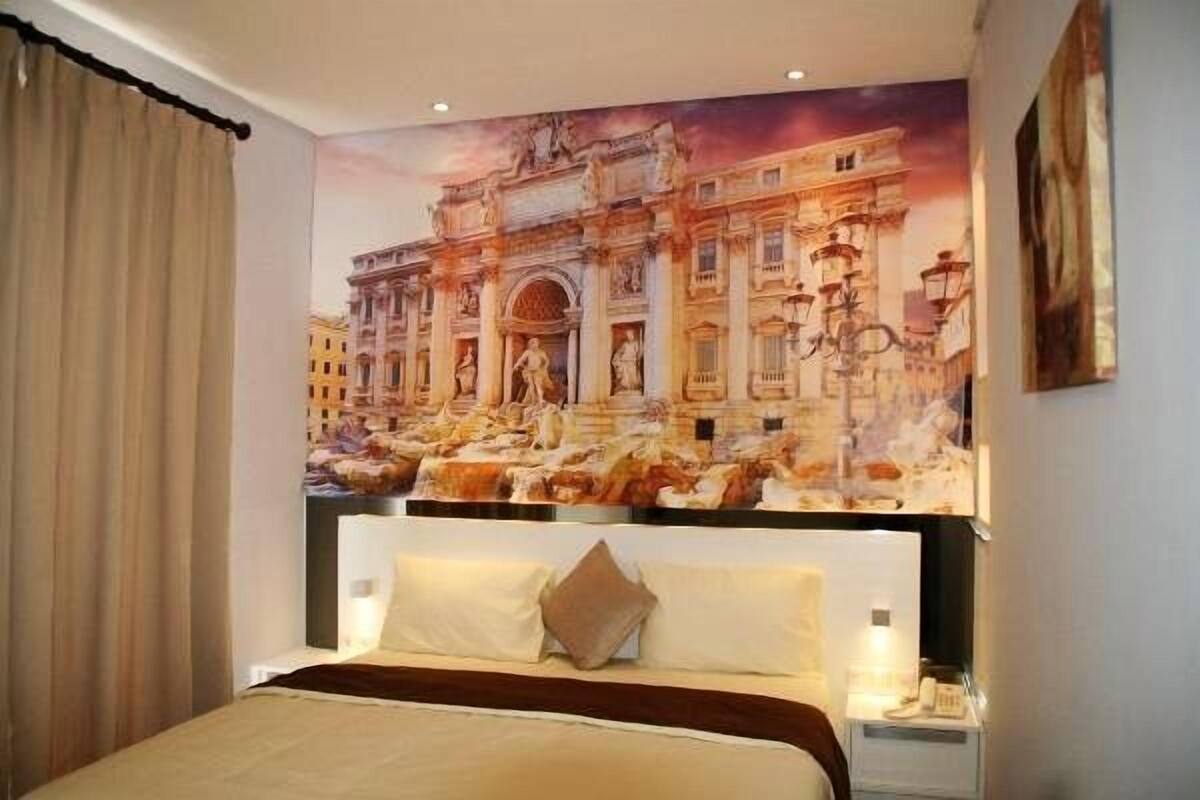 A hotel room with a large bed, a headboard, a nightstand, and a wall mural of a historical building.