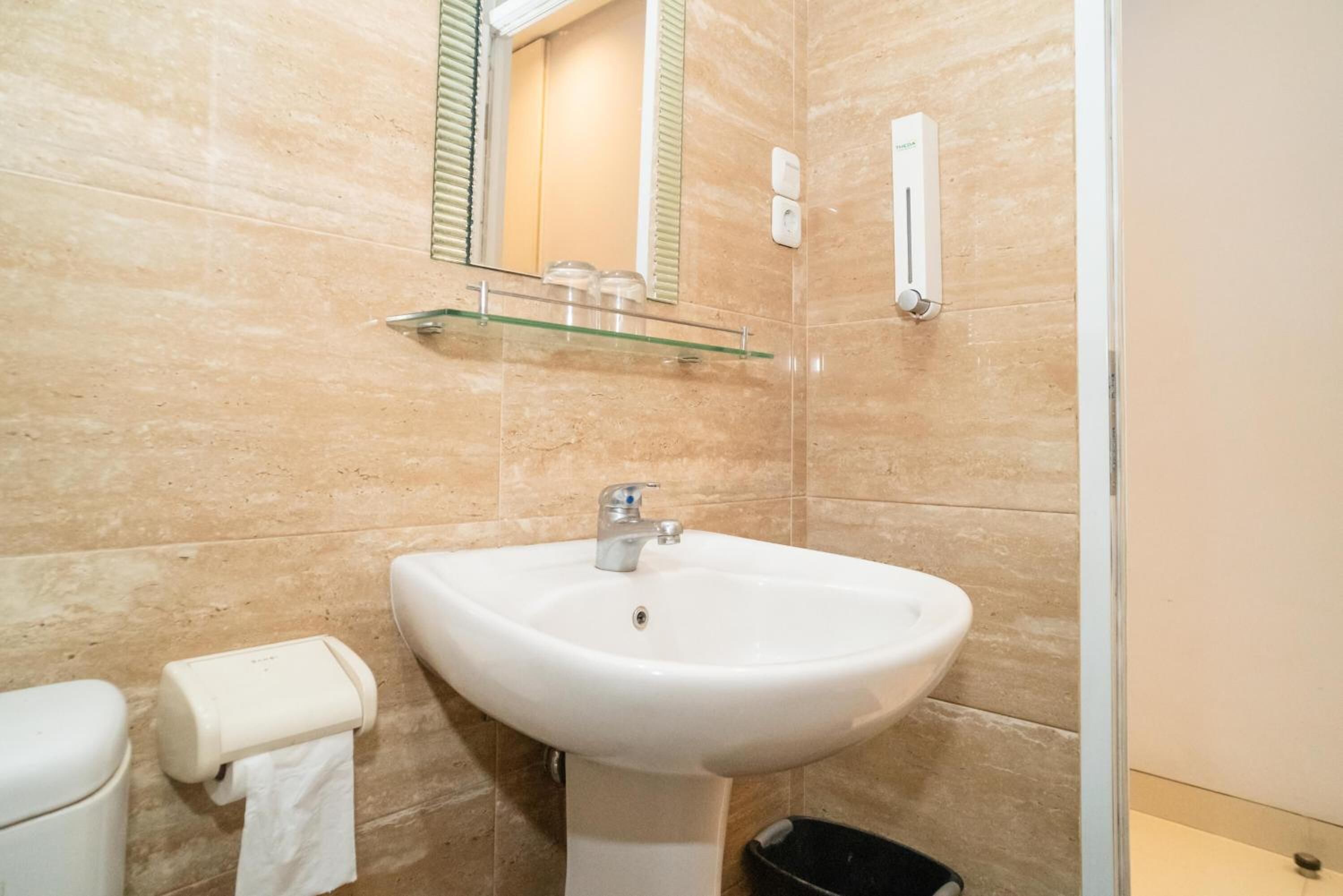 A bathroom with a white sink, a glass shelf above the sink, a toilet with a paper towel holder, and a trash bin.