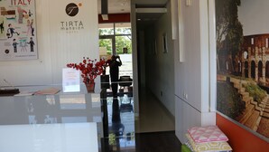 A reception area with a glass counter, a potted plant, a signboard, and a corridor leading to a room with a view of an outdoor structure. - Tirta Mansion Paramount Serpong Hotel (Gading Serpong)