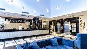A modern reception area with a reception desk, a blue sofa, chandeliers, and a tiled floor. - Prestige Deluxe Hotel & Aquapark Club - All Inclusive (Golden Sands)